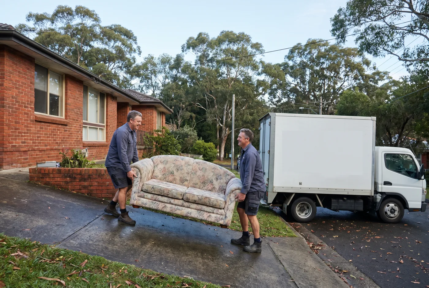 Two crew members carrying an old couch down a sloped driveway toward a box truck