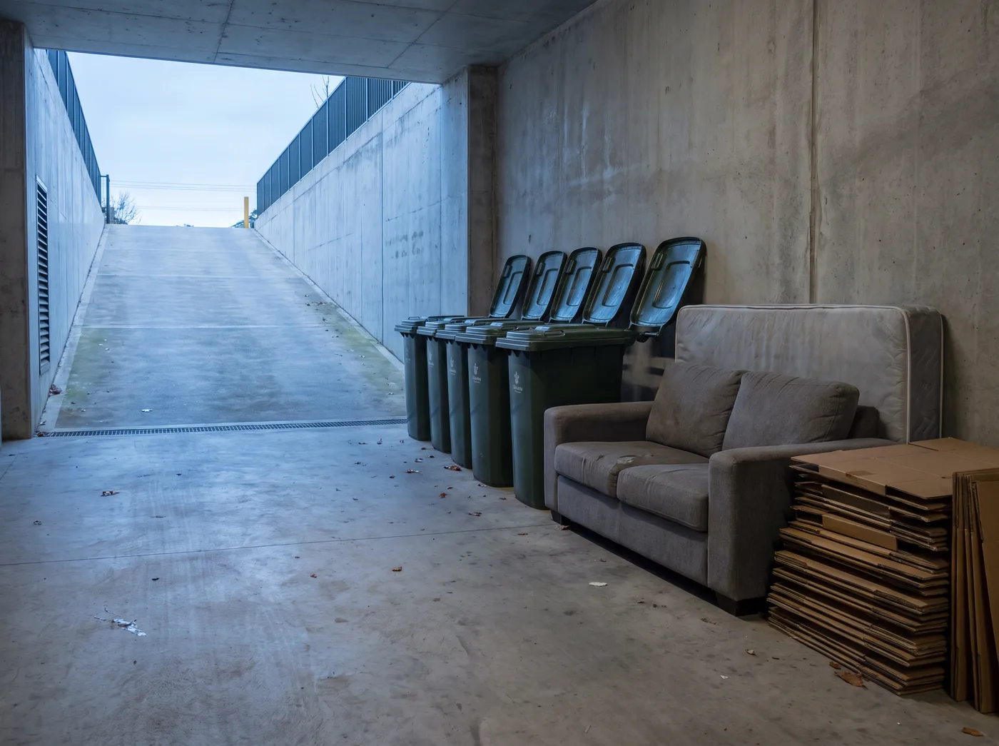 An apartment building bin bay with communal wheelie bins and an abandoned couch, mattress and flattened boxes left beside them