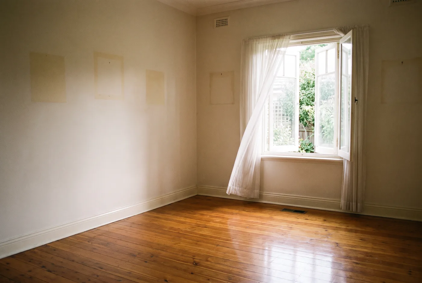 An empty, freshly swept living room in an older home, pale marks on the walls where pictures hung, sheer curtain lifting at an open window over a garden