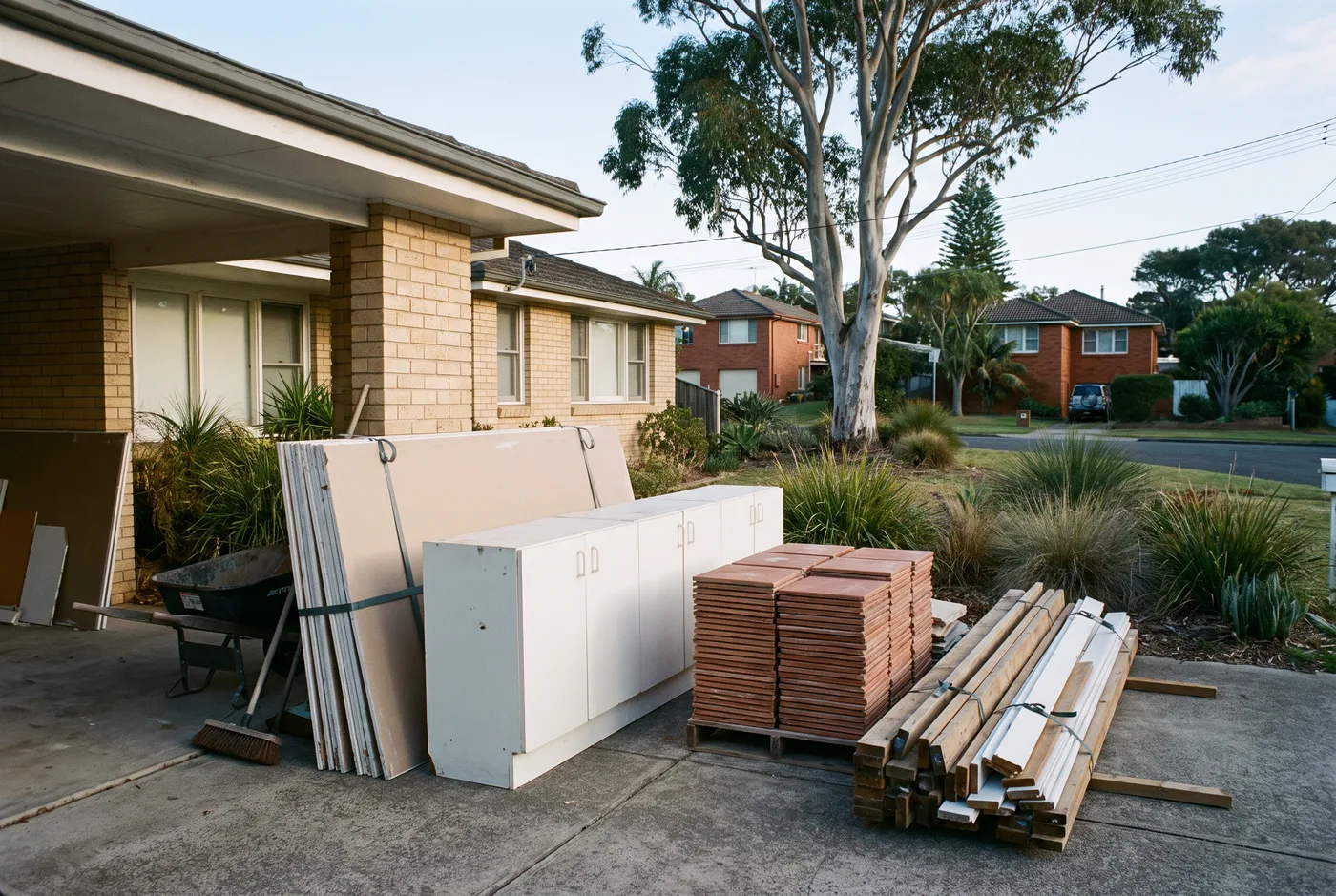 Renovation strip-out debris stacked neatly in an open carport: plasterboard sheets, an old kitchen cupboard run, stacked tiles and bundled timber offcuts