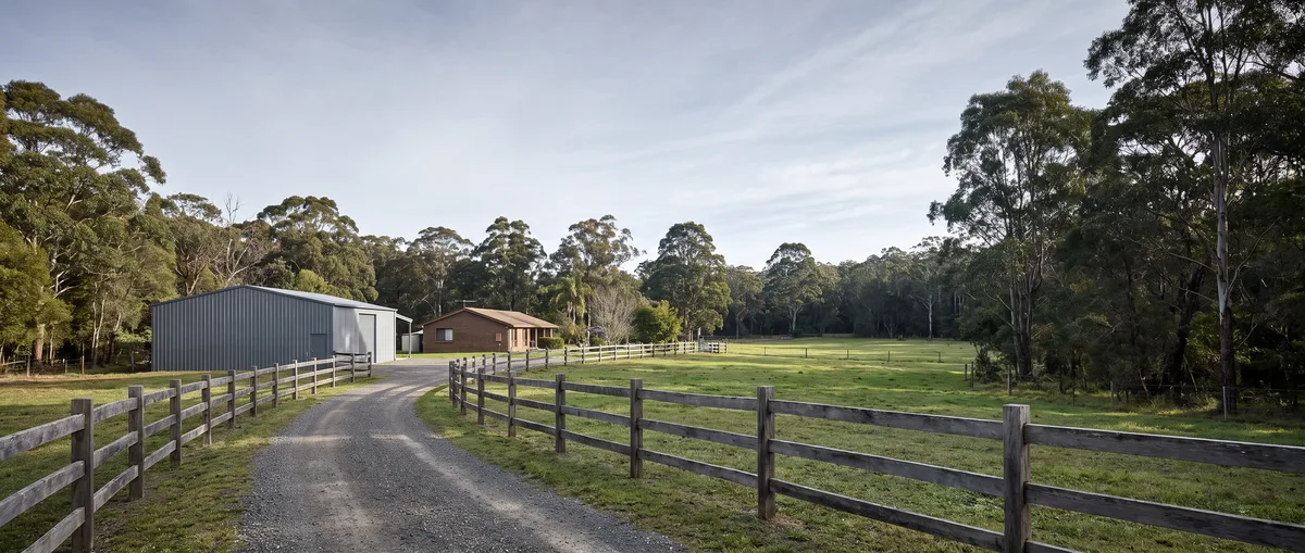 A Terrey Hills acreage property: long gravel driveway past post-and-rail fencing to a large steel machinery shed beside a brick house, open paddock and bushland behind